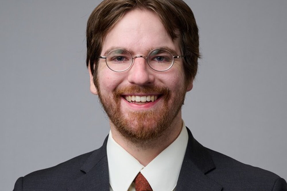 Patrick Aleo. A man wearing a suit with his bangs parted, round glasses and some facial hair.