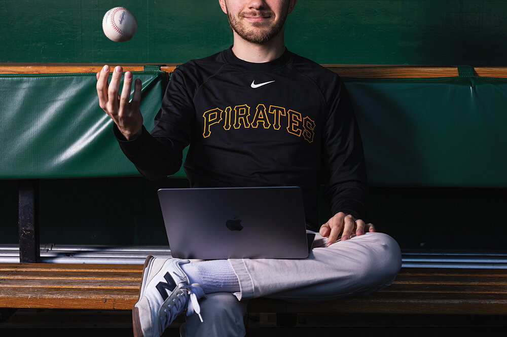 Charlie Young sitting in a dugout with a laptop and tossing a baseball in the air.