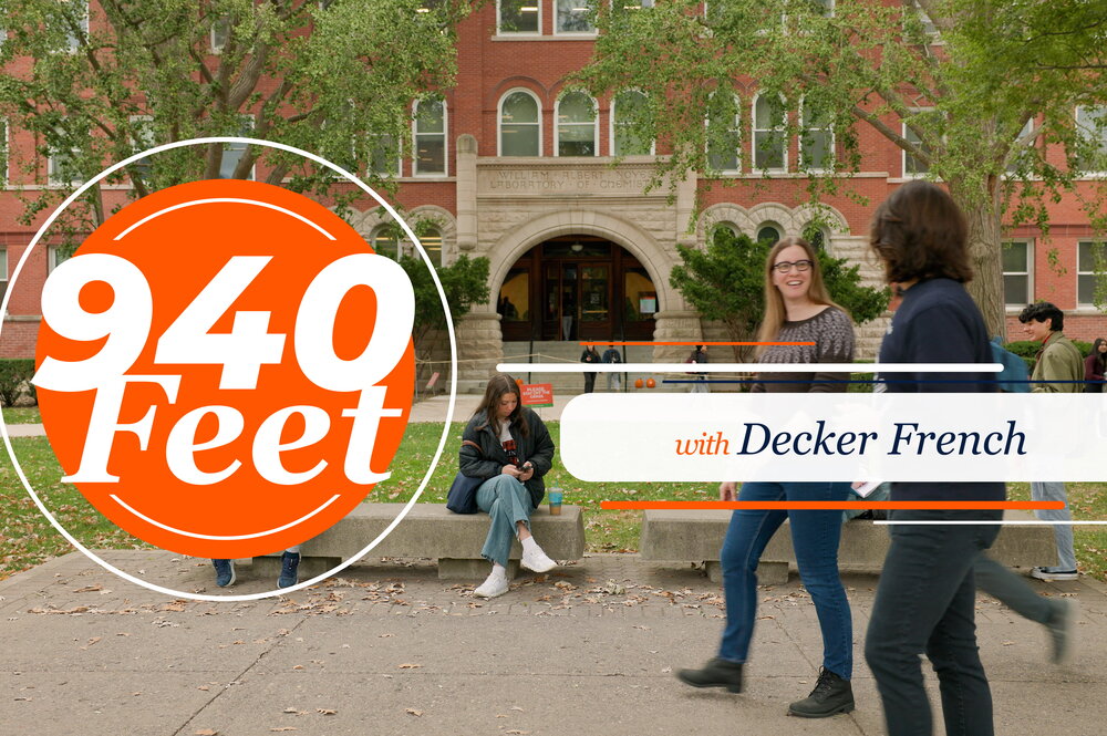 940 Feet with Decker French. Two people walking in front of brick building and people sitting on benches having a conversation.