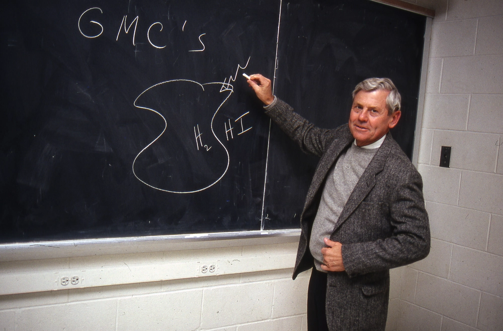 A person writing on a chalkboard while smiling and looking at the camera.
