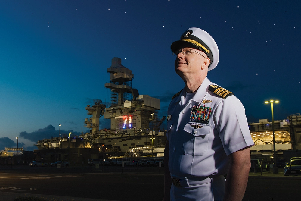 A man in a navy uniform standing in front of a naval ship.