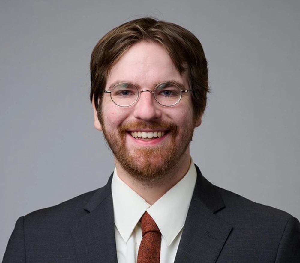 Patrick Aleo. A man wearing a suit with his bangs parted, round glasses and some facial hair.